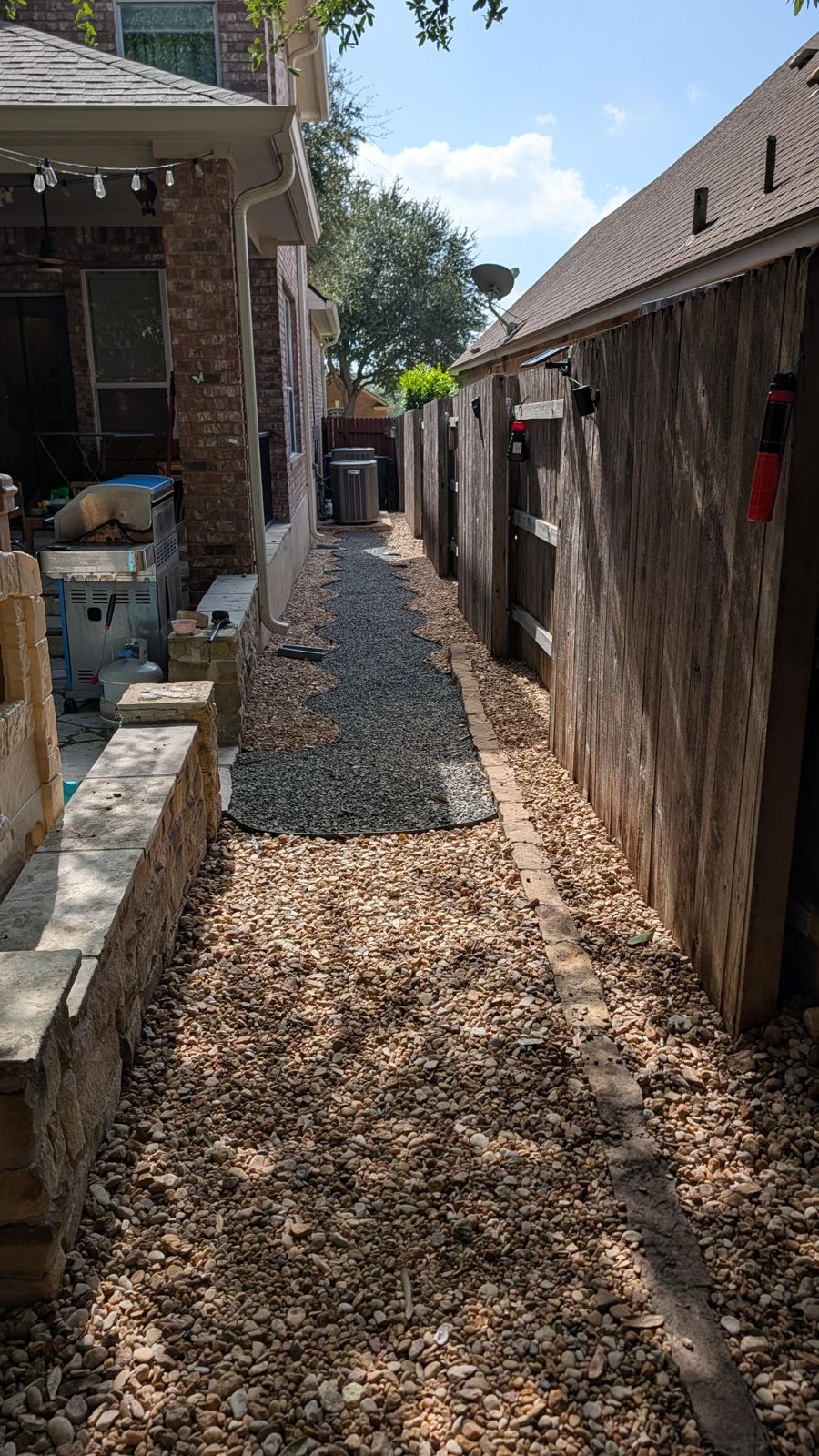 Narrow side yard turned into a curving dark gravel path with steel edging, bordered by river rock and a brick edge along the fence