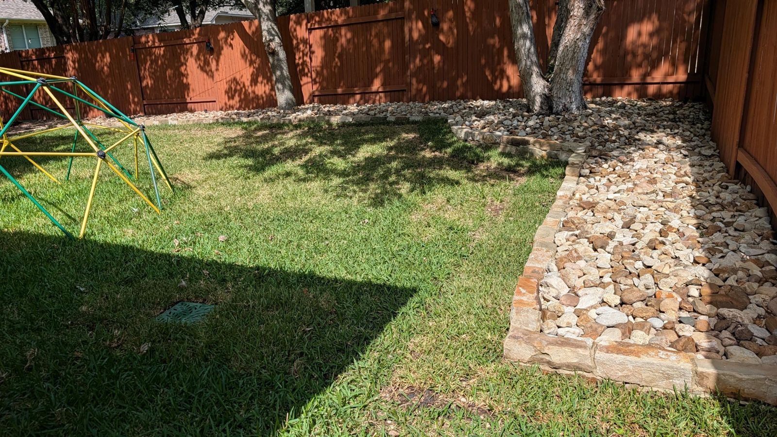 Backyard with a stone-edged river rock bed curving along the fence and around the trees, next to healthy lawn