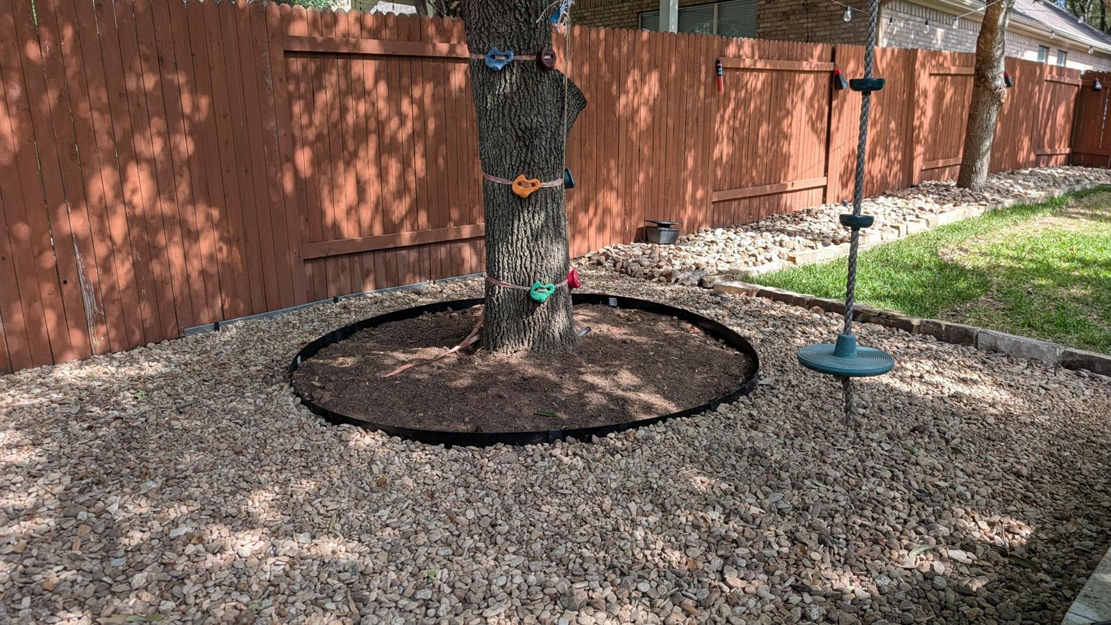 Shade tree with a steel-edged mulch ring surrounded by river rock groundcover, with a stone-edged bed and lawn beyond